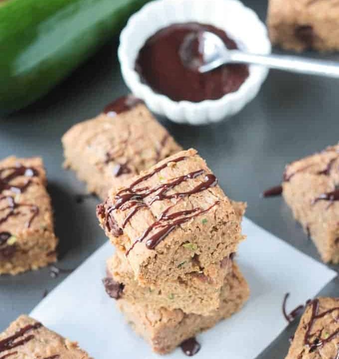 Stack of three chocolate chip zucchini bars, surrounded by more bars and a bowl of chocolate sauce in the background.