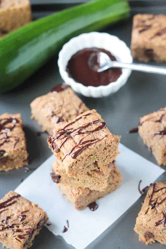 Stack of three chocolate chip zucchini bars, surrounded by more bars and a bowl of chocolate sauce in the background.