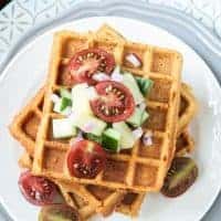 Crunchy corn waffles on a white plate topped with pineapple cucumber salsa and halved cherry tomatoes.