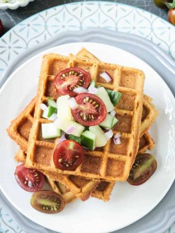 Crunchy corn waffles on a white plate topped with pineapple cucumber salsa and halved cherry tomatoes.