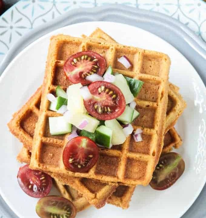 Crunchy corn waffles on a white plate topped with pineapple cucumber salsa and halved cherry tomatoes.