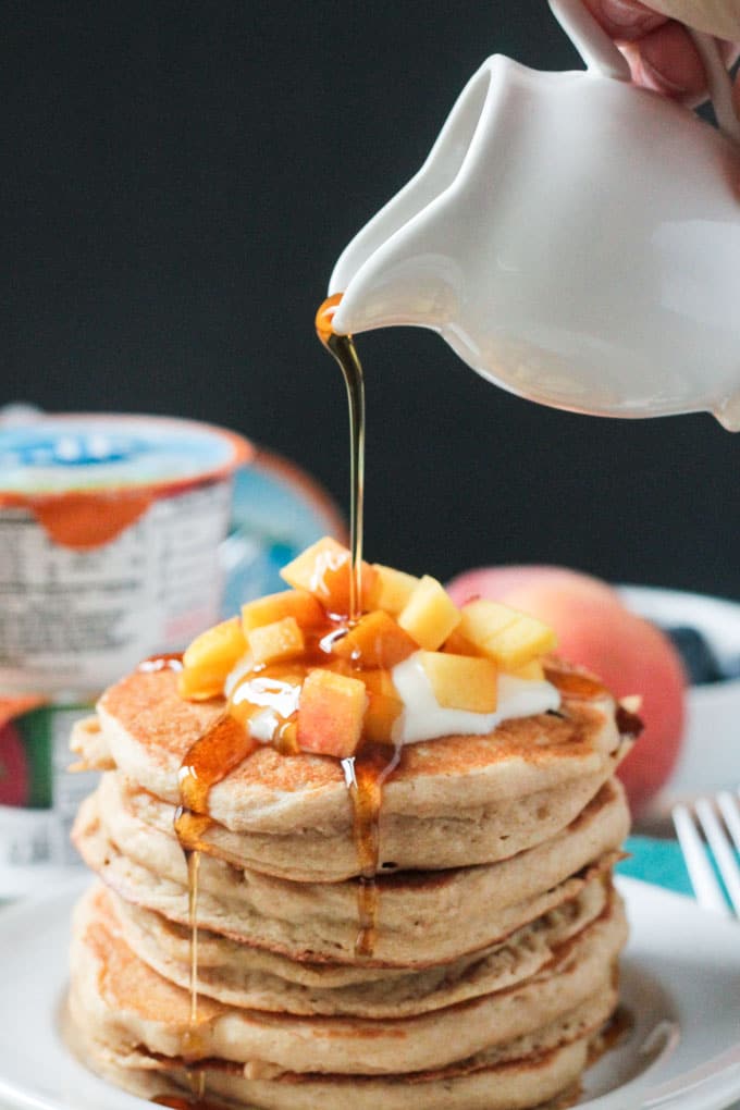 Maple syrup being drizzled over a stack of peach pancakes topped with diced fresh peaches.