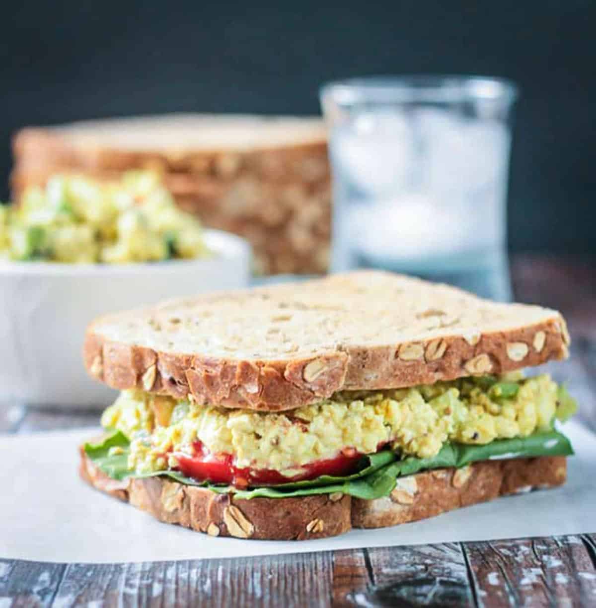 Whole grain bread sandwich with curried tofu salad, lettuce & tomato. Glass of water and a stack of bread in the background.