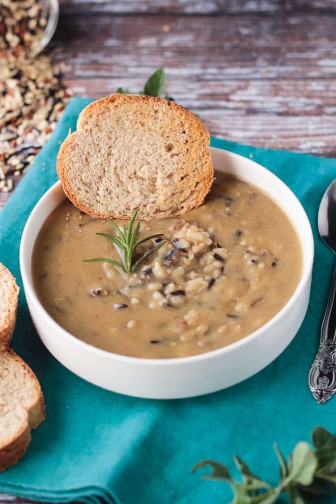 Wild rice soup in a white bowl with a sprig of rosemary and a slice of baguette.