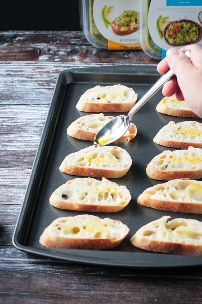 Olive oil being spooned onto slices of baguette on a baking sheet