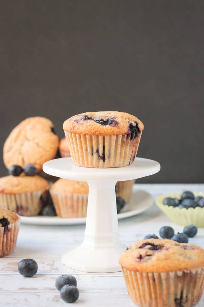 One blueberry muffin on a small cupcake stand in front of a plate off more muffins.