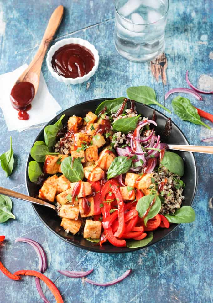 Overhead view of a black bowl filled with baked tofu cubes, roasted red pepper strips, quinoa, fresh spinach, and red onions. One fork in each side of the bowl. Small white bowl of bbq sauce and a small wooden spoon with bbq sauce on it behind.