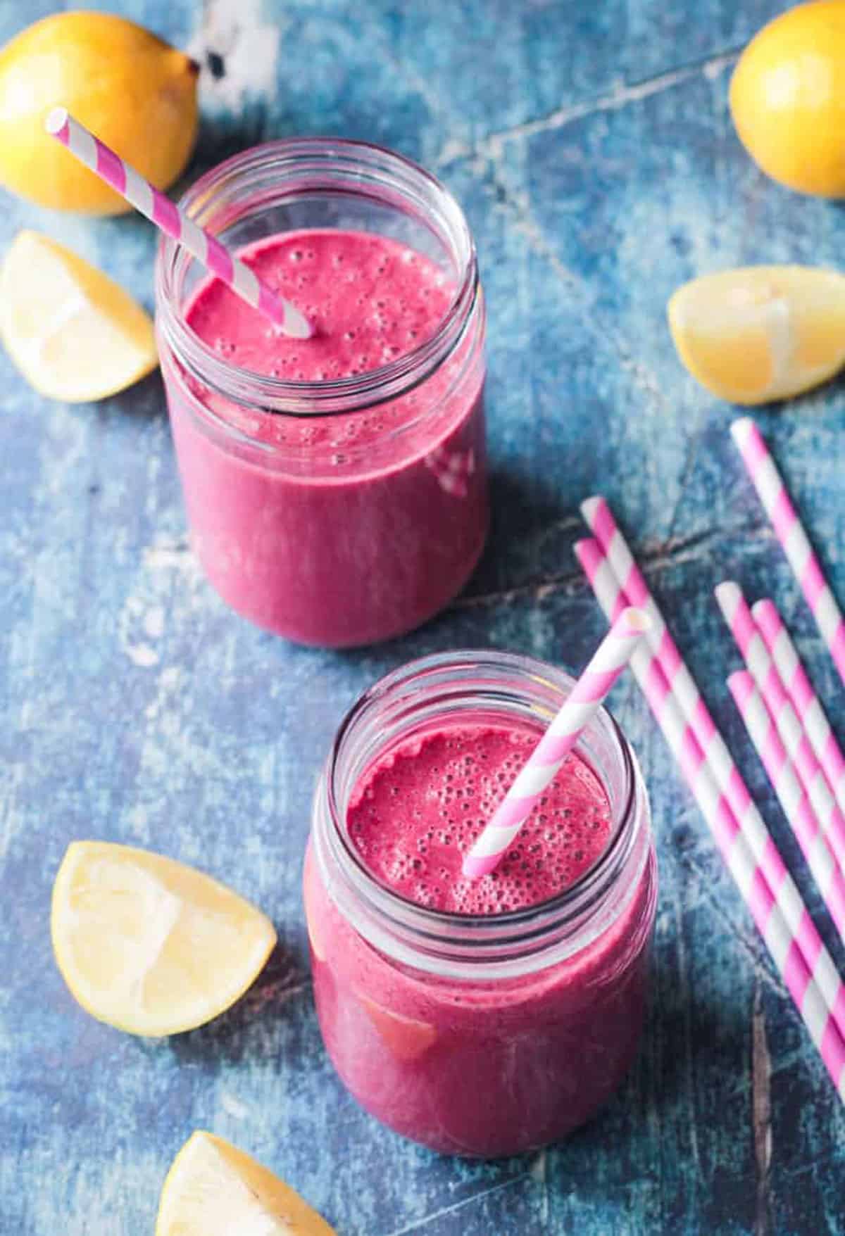 Strawberry beet smoothies in glass jars with pink and white straws on a blue background.