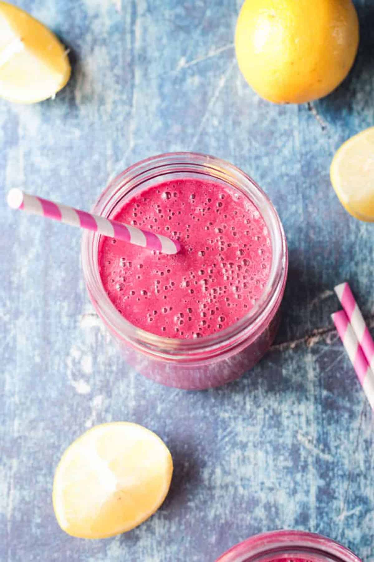 Overhead view of a bright pink Strawberry Beet Smoothie in a glass jar on a dark blue slate board with a pink and white striped straw. Two lemon wedges lie nearby.