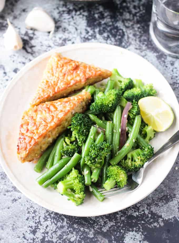 White plate with green bean broccoli stir fry with two slices of tempeh, a lemon wedge, and a fork resting on the side.