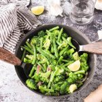 Two wooden handled serving spoons in either side of a skillet with green bean broccoli stir fry. Gray and white striped dish towel, lemon wedges, and garlic slices nearby.