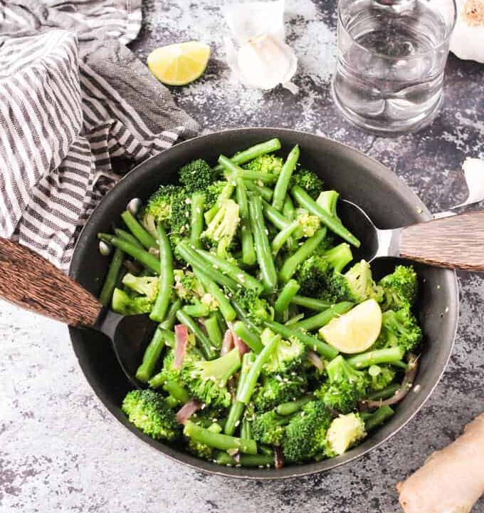 Two wooden handled serving spoons in either side of a skillet with green bean broccoli stir fry. Gray and white striped dish towel, lemon wedges, and garlic slices nearby.