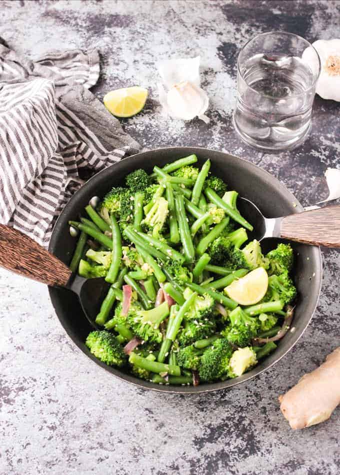 Two wooden handled serving spoons in either side of a skillet with green bean broccoli stir fry. Gray and white striped dish towel, lemon wedges, and garlic slices nearby.