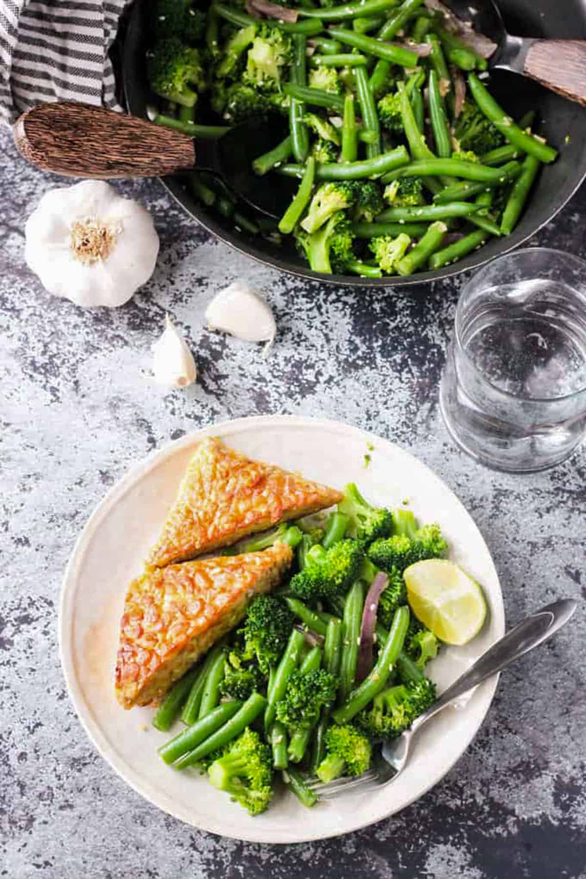 Easy Green Bean Broccoli Stir Fry. An easy weeknight vegetable side dish that takes less than 30 minutes. Just a few ingredients are needed, but it brings lots of flavor and nutrients. Serve it as a side or over pasta or rice. Broccoli stir fry on a plate with tempeh triangles and a skillet of stir fry behind.