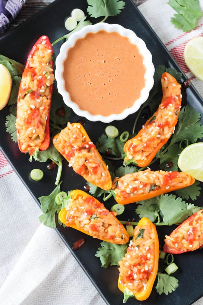 Black rectangular plate of spicy peanut rice stuffed mini peppers and a bowl of peanut sauce.