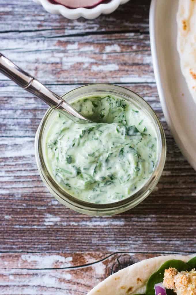 Metal spoon dipped into a small glass jar of cilantro yogurt sauce.