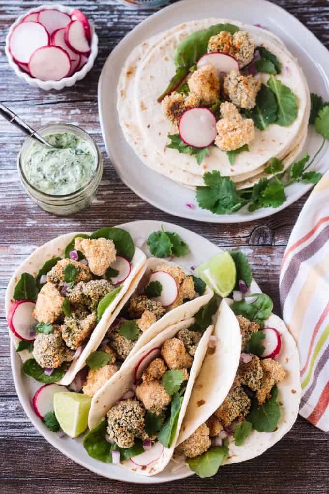 Overhead view of a plate of Crunchy Veggie Tacos filled with breaded cauliflower and broccoli, sliced radishes, greens, and garnished with lime wedges. Cilantro yogurt sauce in a small jar behind next to a plate of tortillas.