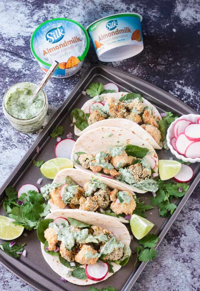 Four Crunchy Veggie Tacos on a baking sheet topped with Cilantro Yogurt Sauce. Two containers of Silk Plain Almond Dairy-Free Yogurt Alternative in the background. 