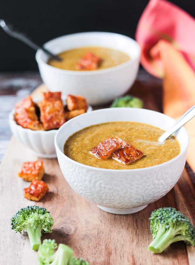 Two bowls of vegan broccoli soup with metal spoons in them on a wooden board.
