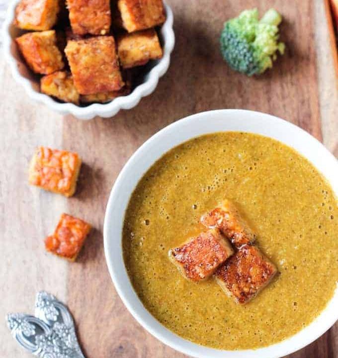 Overhead view of a bowl of vegan broccoli soup topped with three tempeh "croutons." More croutons and a fresh broccoli floret lie nearby.