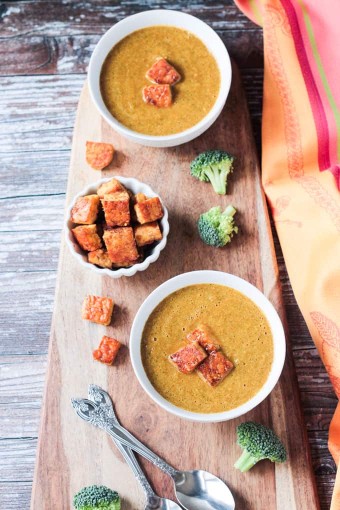 Overhead view of two bowls of vegan broccoli soup. Small bowl of crispy tempeh croutons in the middle.