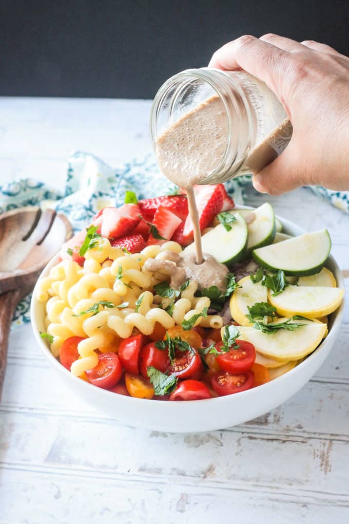 Jar of creamy balsamic dressing being poured over a bowl of summer pastas salad with fresh veggies and strawberries.
