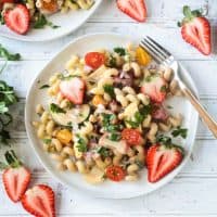 Summer pasta salad on a white plate on a wide wooden picnic table. Wooden handled fork on the side of the plate. Fresh halved strawberries on the table around the plate.