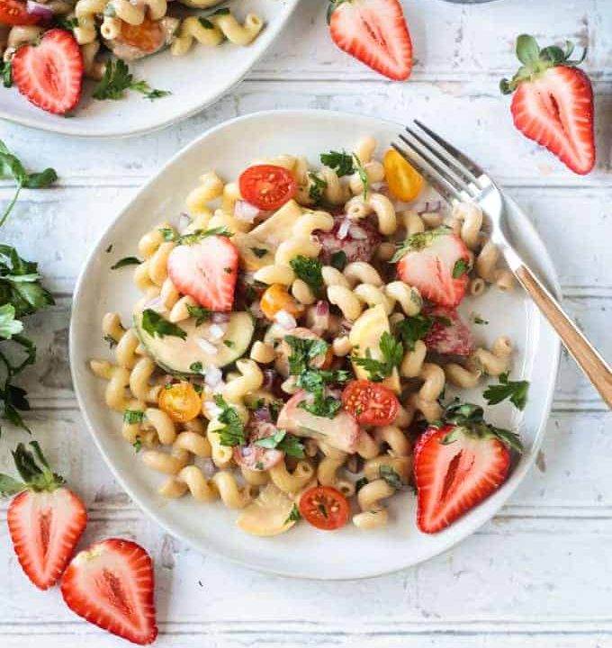 Summer pasta salad on a white plate on a wide wooden picnic table. Wooden handled fork on the side of the plate. Fresh halved strawberries on the table around the plate.