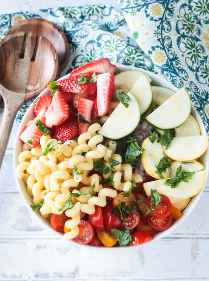 Bowl of summer pasta salad ingredients, including curly pasta, sliced strawberries, zucchini, summer squash, cherry tomatoes, and fresh parsley.