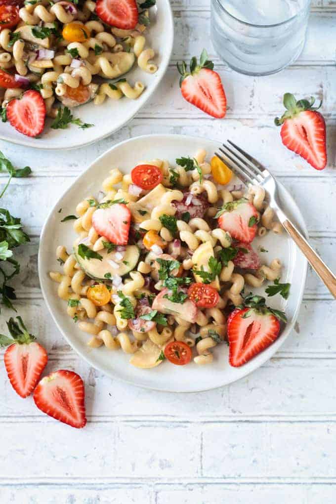 Summer pasta salad on a white plate on a wide wooden picnic table. Wooden handled fork on the side of the plate. Fresh halved strawberries on the table around the plate.