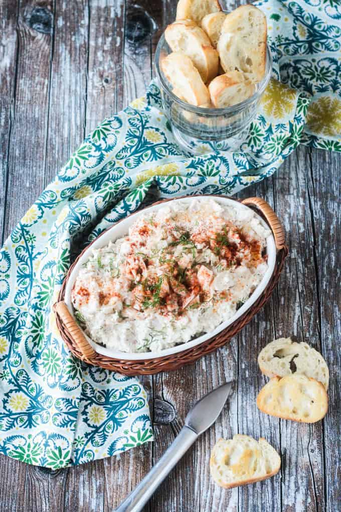 Overhead view of a bowl of cold crab dip on a blue/green dish towel. Baguette slices on the table nearby.