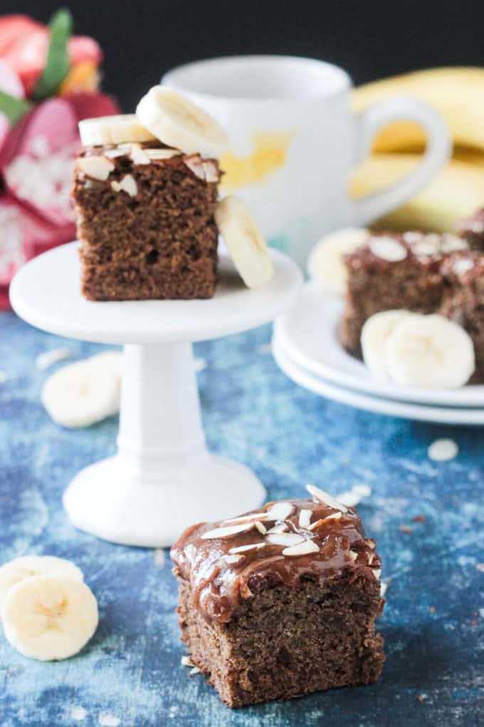 One slice of banana bread on a blue table. Behind one slice of cake sits on a small cupcake stand. Behind that a place of more cake.