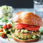 Front view of a falafel burger on a pretzel bun with sliced tomatoes and cucumber peanut relish.