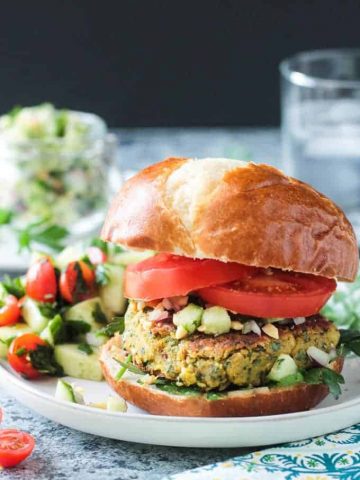 Front view of a falafel burger on a pretzel bun with sliced tomatoes and cucumber peanut relish.