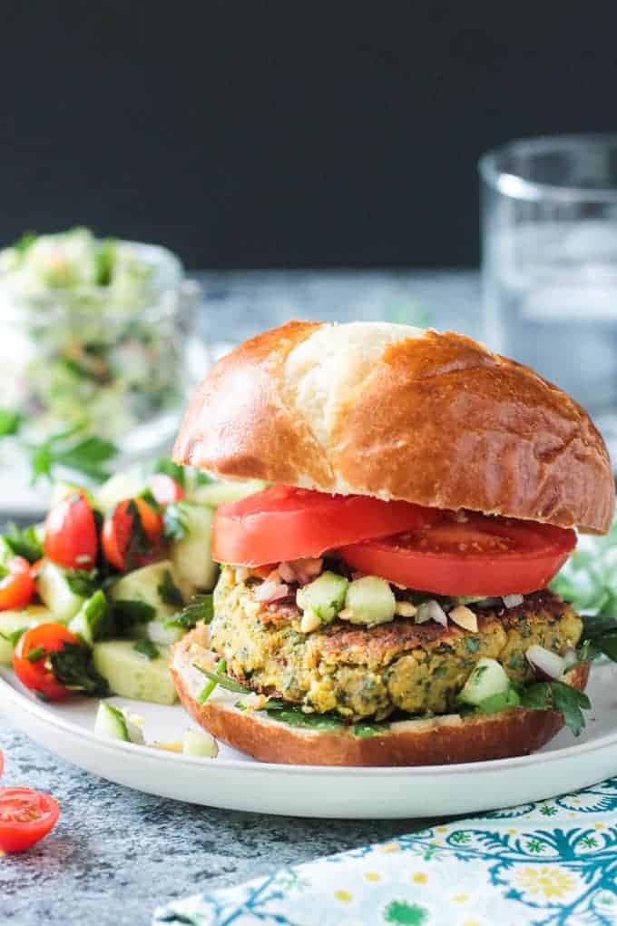 Front view of a falafel burger on a pretzel bun with sliced tomatoes and cucumber peanut relish.