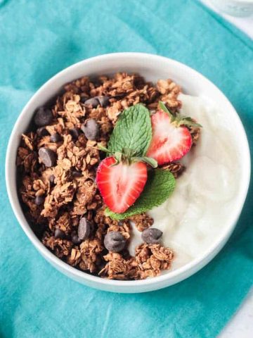 Bowl of healthy homemade granola with chocolate chips, vanilla yogurt, and fresh sliced strawberries and mint leaves.