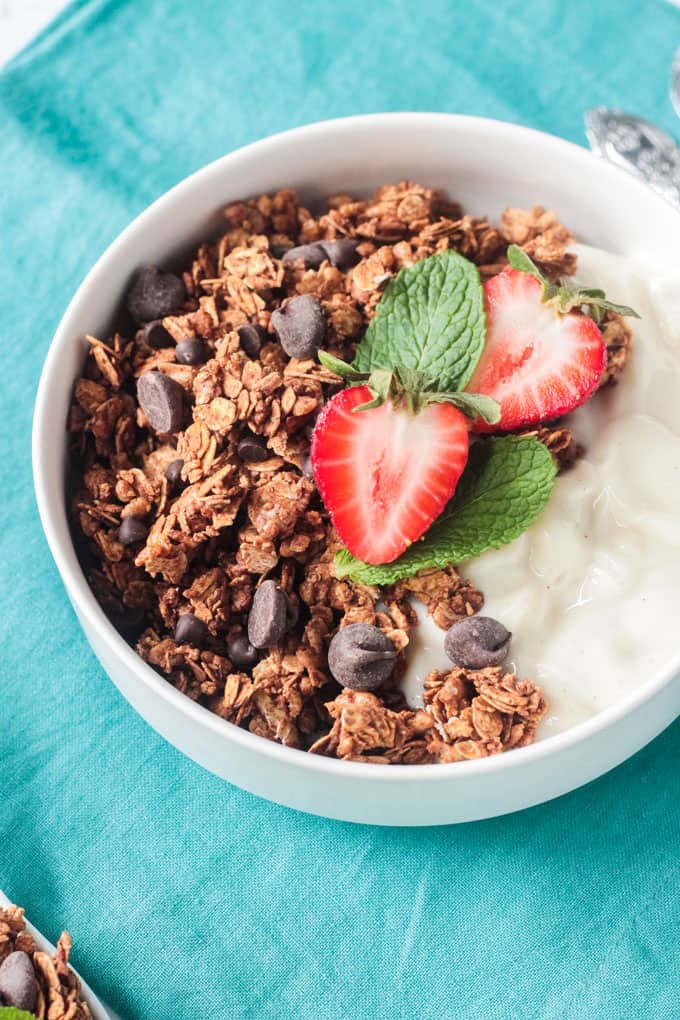 Close up of crunchy Mint Chocolate Chip Healthy Homemade Granola in a white bowl with yogurt and topped with halved strawberries and mint leaves.