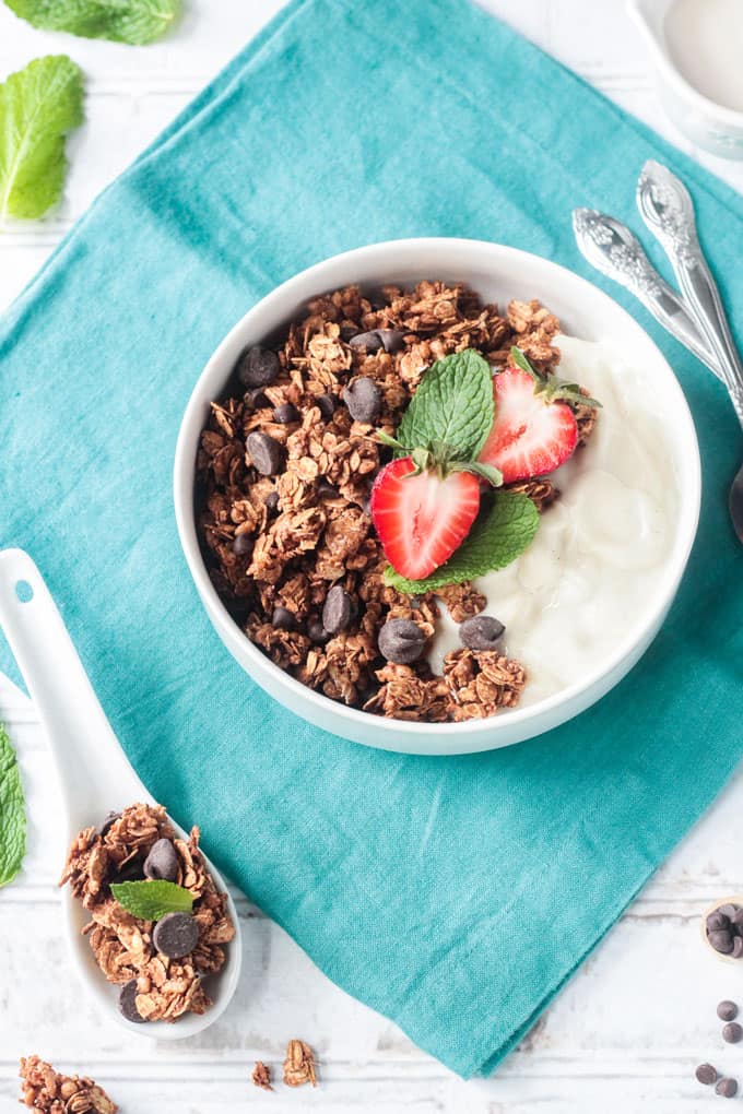 Bowl of yogurt and granola on a blue napkin next to a white spoon holding a scoop of granola.