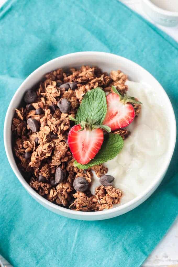 Bowl of healthy homemade granola with chocolate chips, vanilla yogurt, and fresh sliced strawberries and mint leaves.