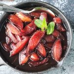 Close up overhead view of poached nectarines in red wine, topped with mint sprigs and cloves, in a cast iron skillet.