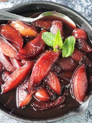 Close up overhead view of poached nectarines in red wine, topped with mint sprigs and cloves, in a cast iron skillet.