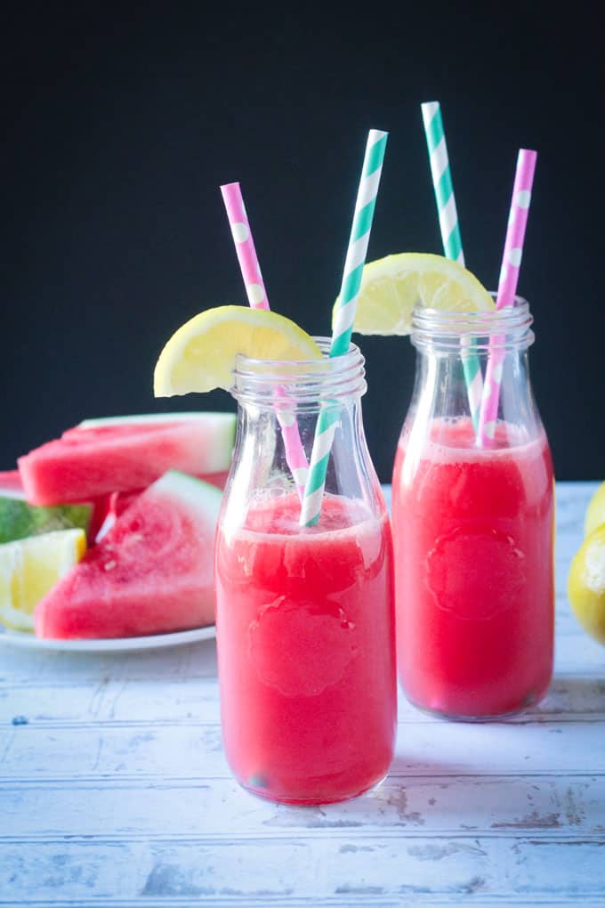 Two glasses full of watermelon juice. One blue/white striped straw and one pink/white striped straw in each glass. Plate of sliced watermelon behind.