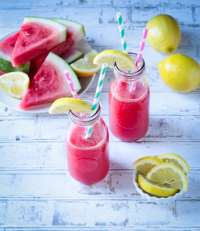 Overhead view of two glasses of watermelon juice next to a plate of watermelon slices and a small bowl of lemon wedges.