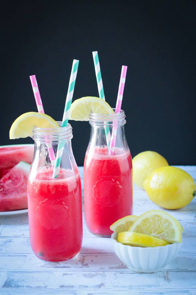 Two glasses of watermelon juice with a small white bowl of lemon slices on the table nearby. Two whole lemons behind.