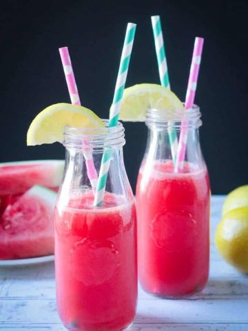 Two glass bottles of watermelon juice with straws and lemon wedges on top.