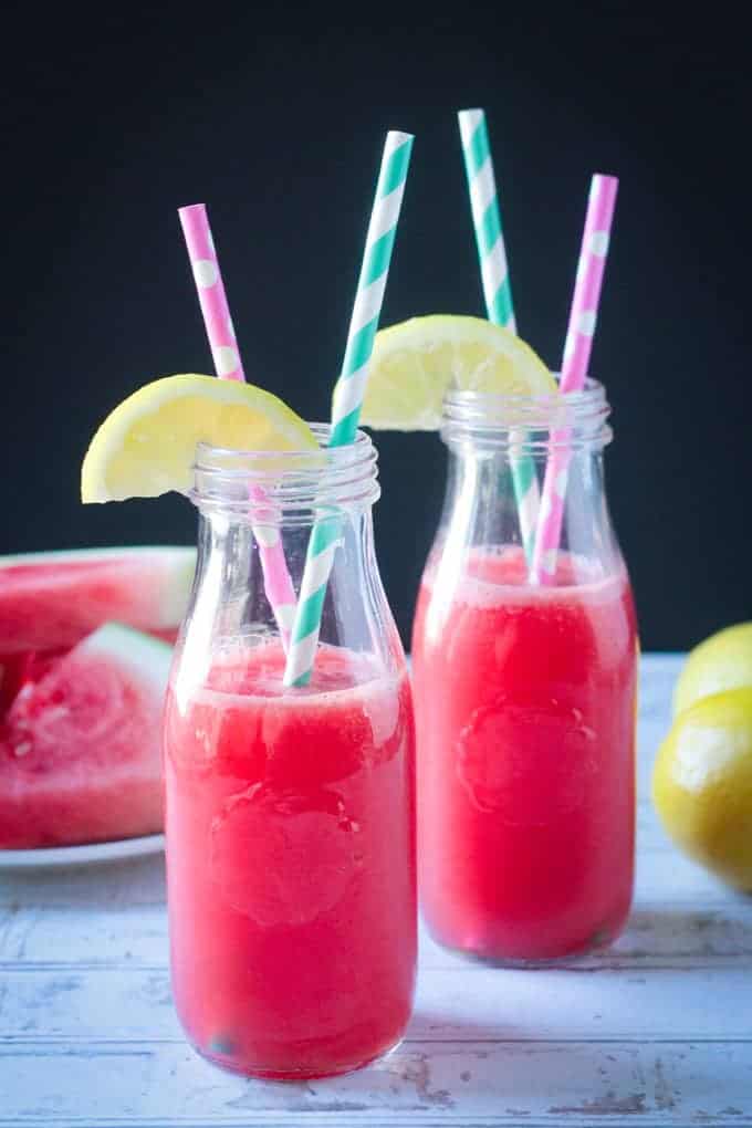 Two glass bottles of watermelon juice with straws and lemon wedges on top.