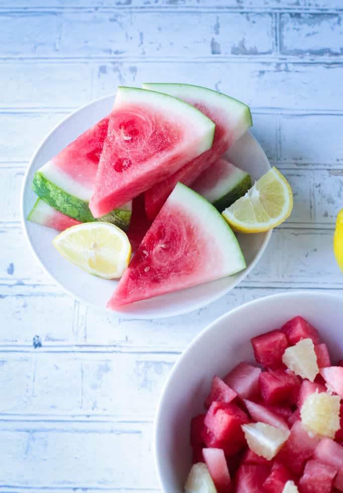 A pile of watermelon slices on a white plate. Two lemon wedges amongst the watermelon.