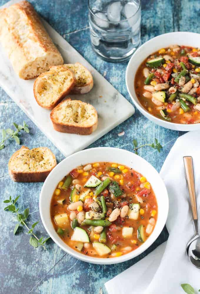 White Bean Soup recipe in a white bowl next to a white napkin with a wooden handled spoon. Loaf of sliced crunchy bread on the table behind next to another bowl of soup.