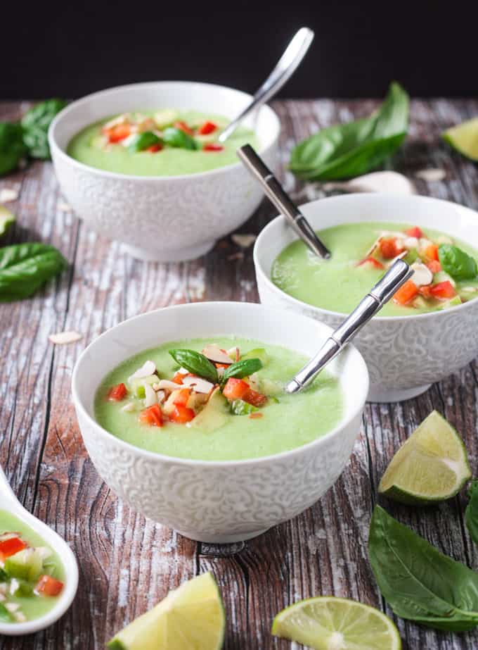 Front view of a bowl of cold cucumber soup garnished with diced red peppers and cucumbers, chopped almonds, and a tiny basil leaf.