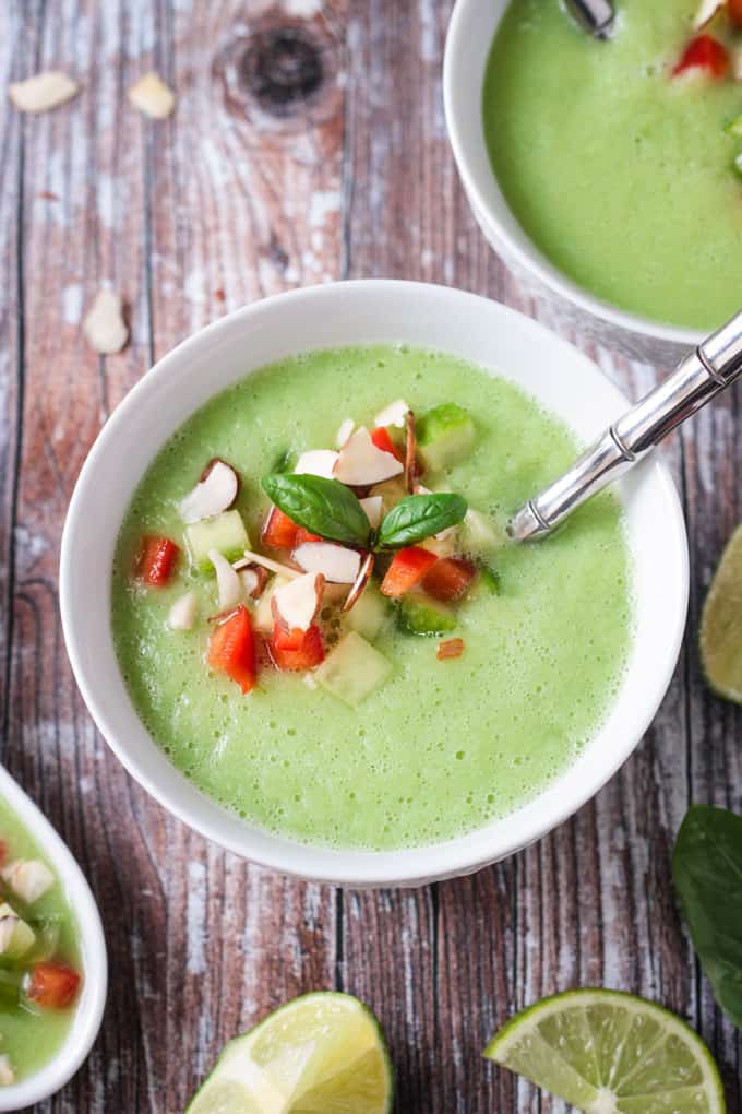 Close up overhead view of cold cucumber soup in a white bowl topped with chopped almonds, diced red pepper, and diced cucumber. A silver metal spoon is in the bowl.